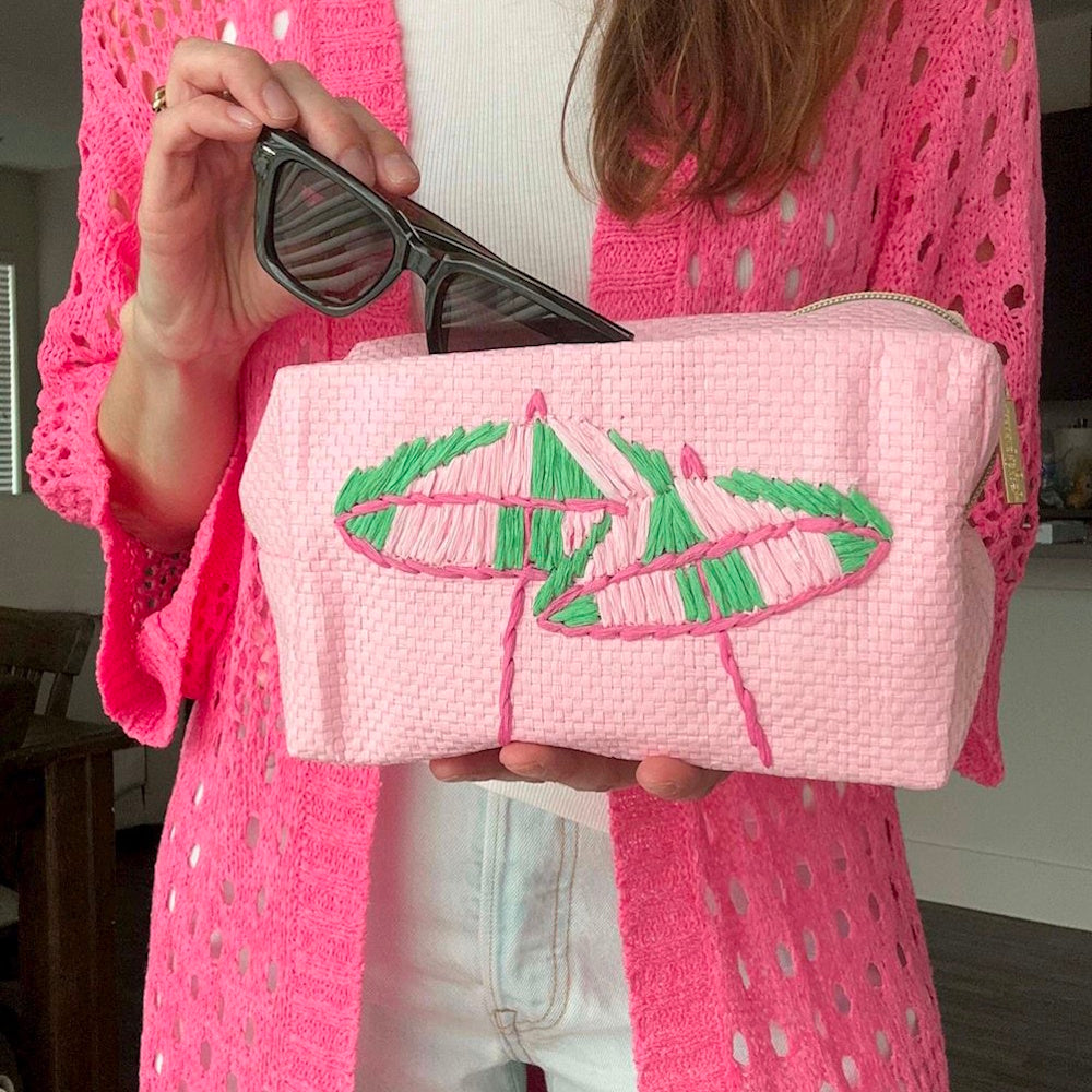 A person holding a pink woven pouch with embroidered green and pink umbrellas, putting sunglasses inside.