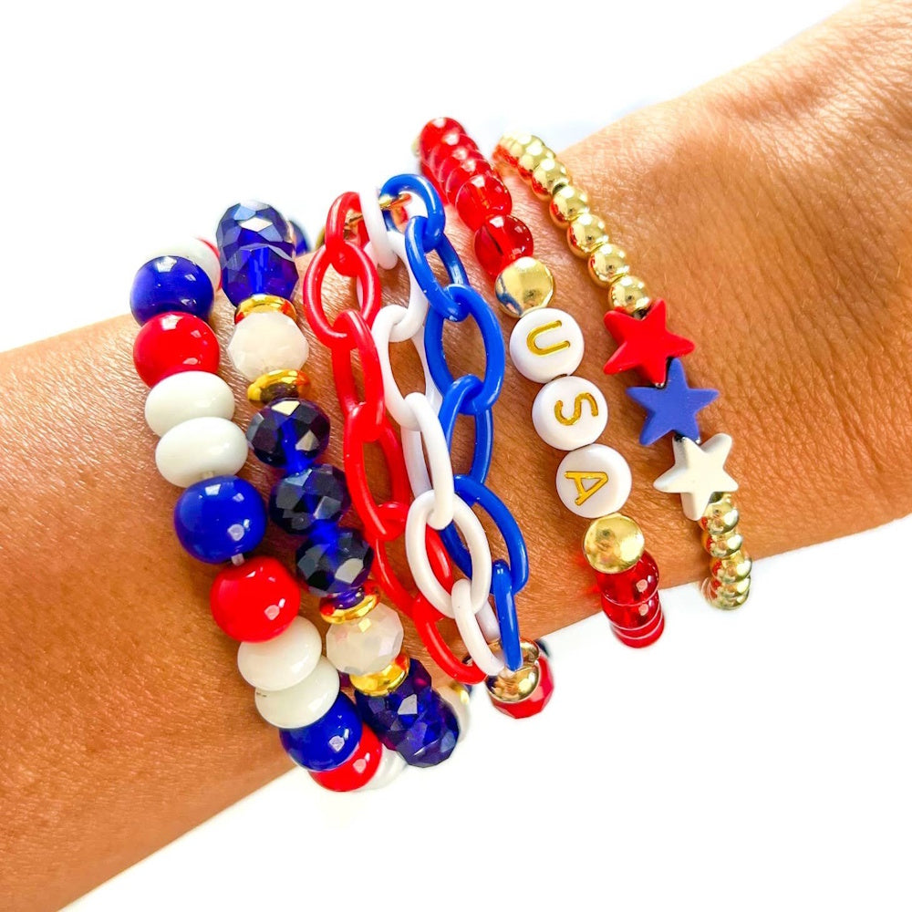 A red, white, and blue 5-piece bracelet stack on a wrist, featuring stars and beads that spell "USA."