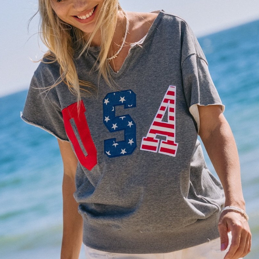 Smiling woman wearing a gray French terry top with "USA" in red, white, and blue letters at the beach.