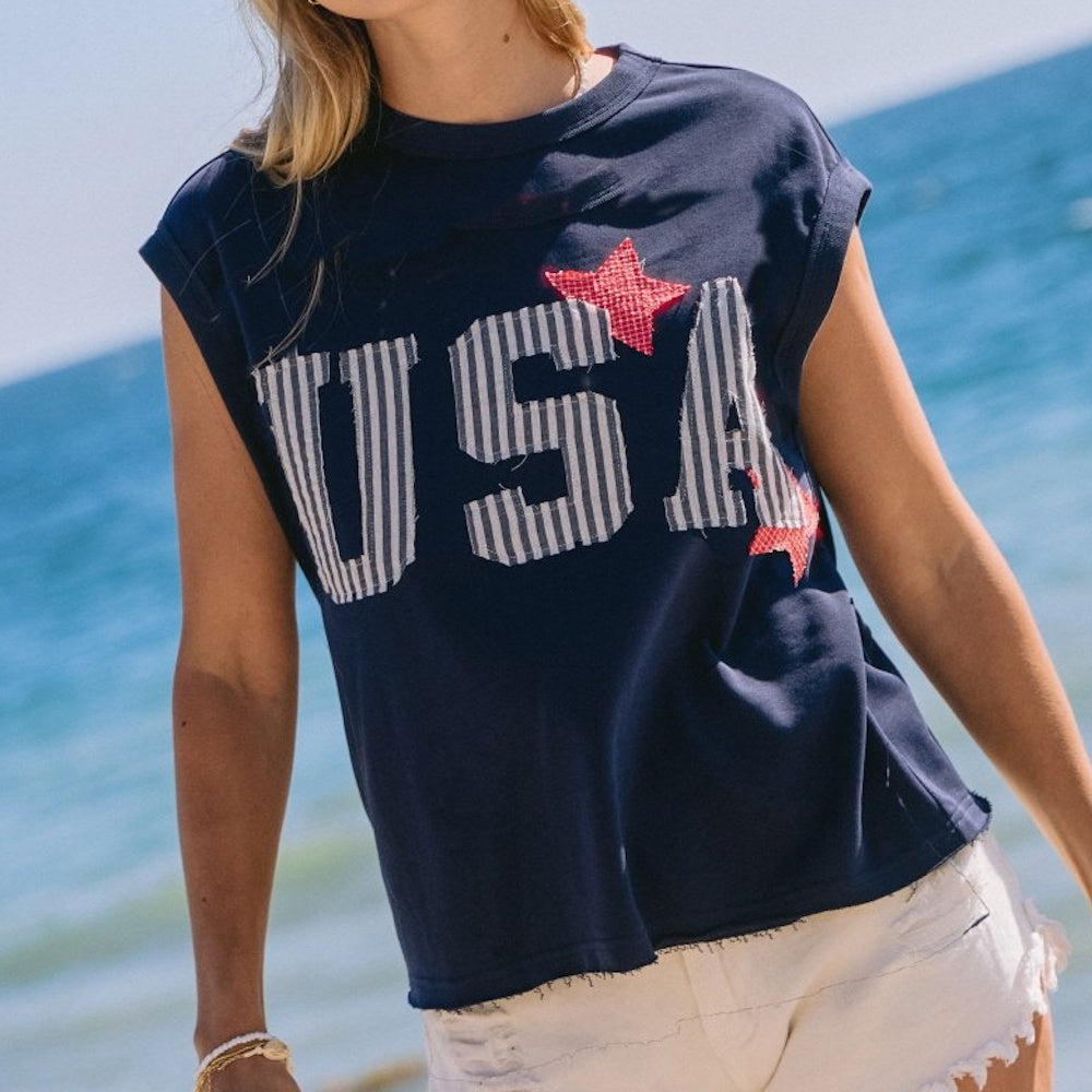 A person wearing a navy muscle tank top with striped "USA" lettering and red stars at the beach.