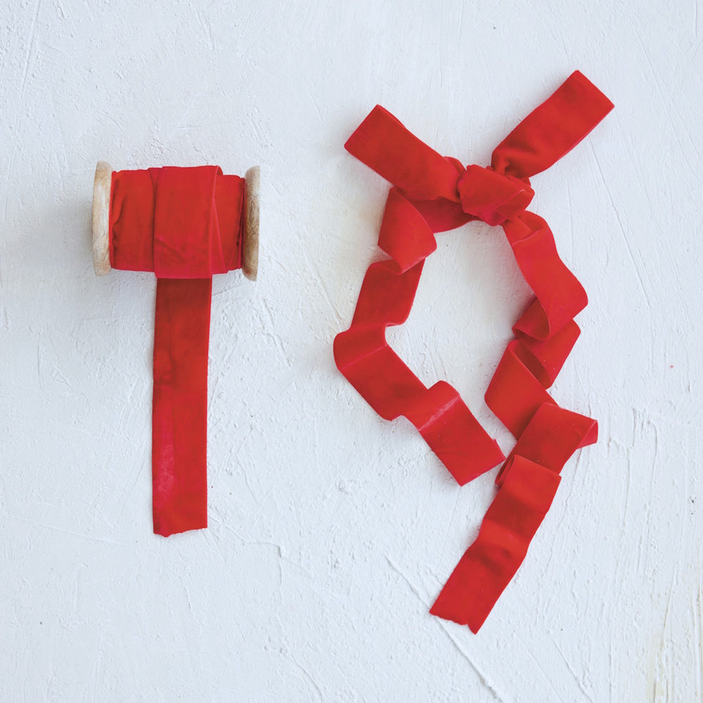 Red velvet ribbon wound on a wooden spool alongside a loosely tied ribbon bow on a white surface.