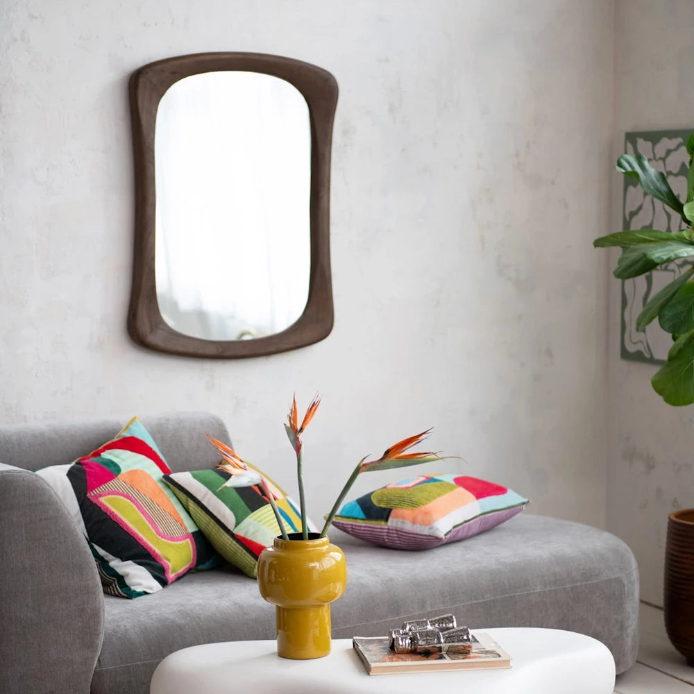 Mid-century modern wall mirror with a curved walnut mango wood frame hanging above a grey sofa with colorful pillows.