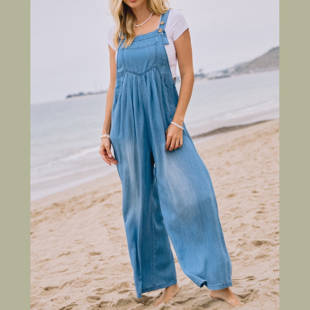 A woman standing on a beach wearing a light blue chambray overall jumpsuit over a white t-shirt.