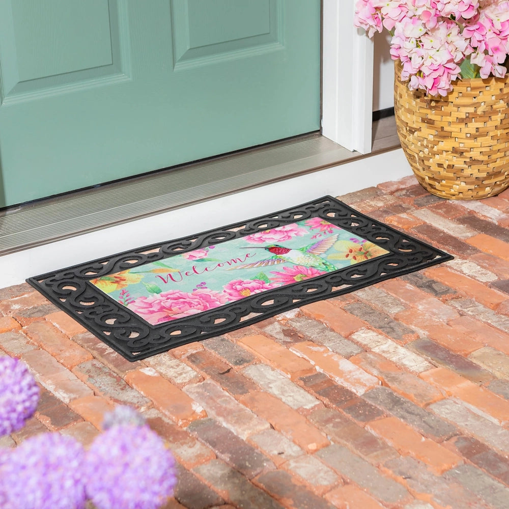 Floral hummingbird welcome doormat with pink flowers on a brick porch, featuring a decorative black scrollwork border.