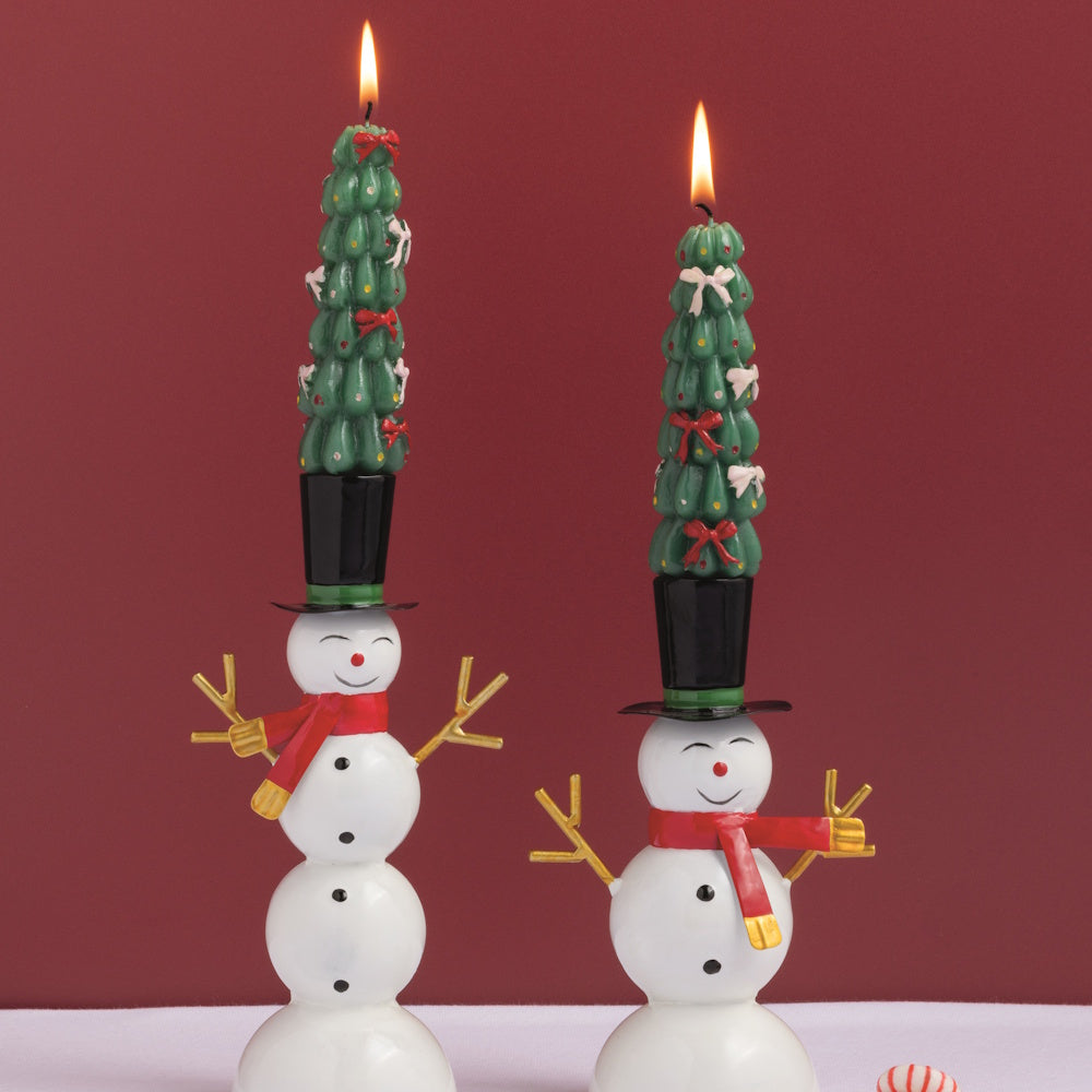 Front view of two lit green wax Christmas tree mini taper candles placed inside snowman candle holders on a table.