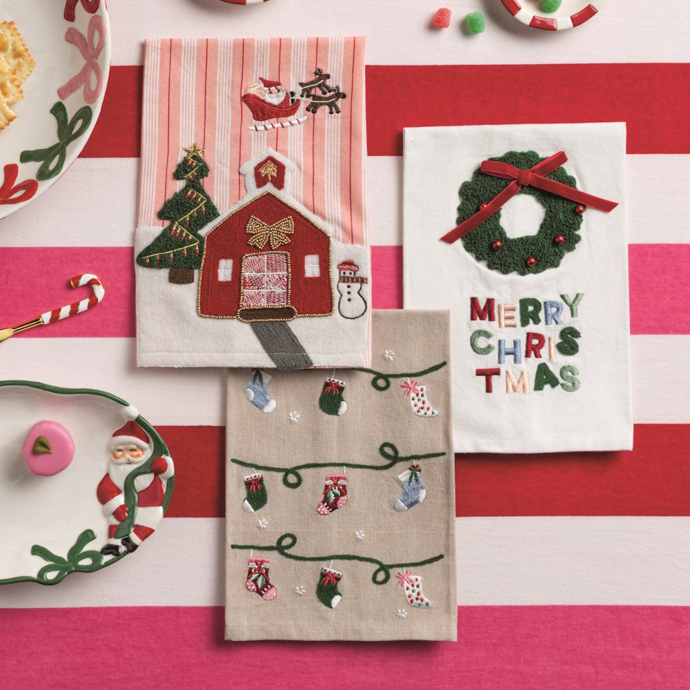Set of three Christmas kitchen towels displayed on a red and white striped background alongside holiday tableware.