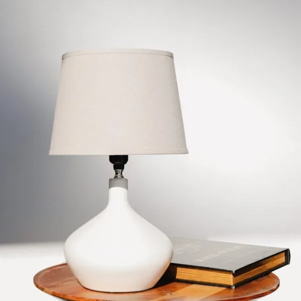 Modern white ceramic table lamp with a tapered linen shade sitting on a wooden side table next to a black book.