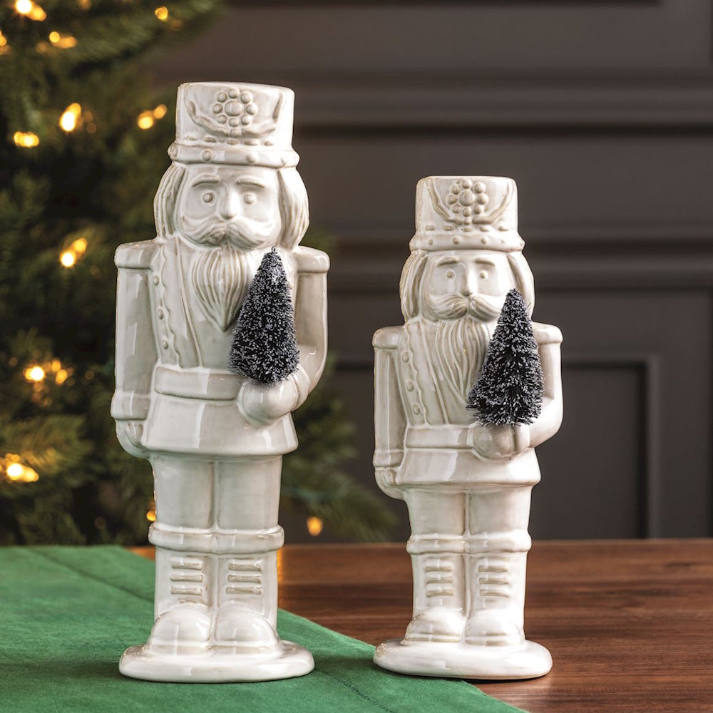 Two white glazed ceramic nutcrackers in different sizes holding small dark frosted bottle brush trees.