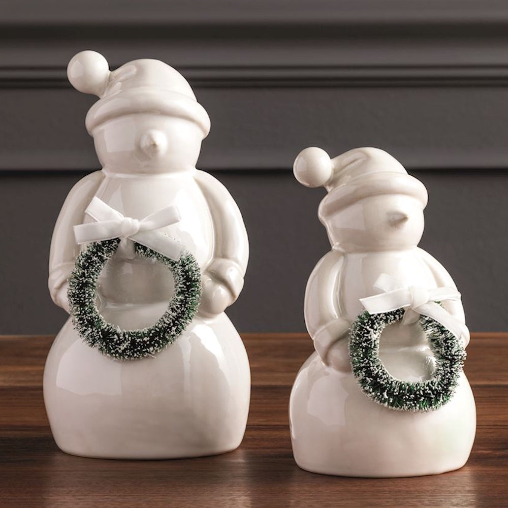 Two white ceramic snowman figurines wearing winter hats and holding miniature pine wreaths with bows.
