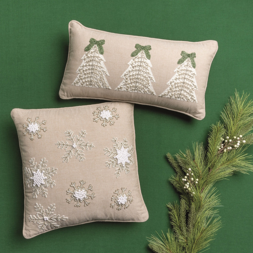 Beige pillows with white beaded Christmas trees and snowflakes, displayed on a green background with pine branches.