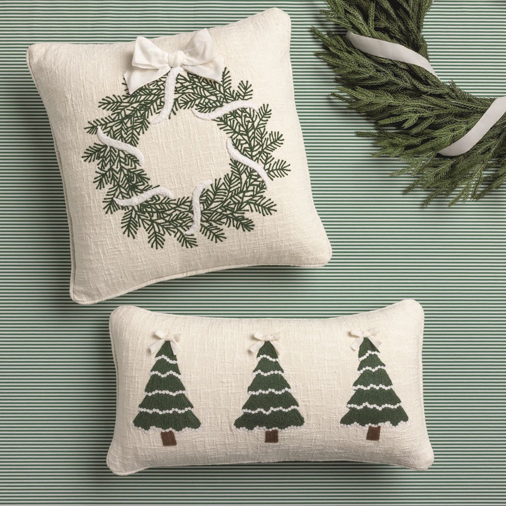 Square and lumbar embroidered holiday pillows displayed with an evergreen wreath on a green striped backdrop.