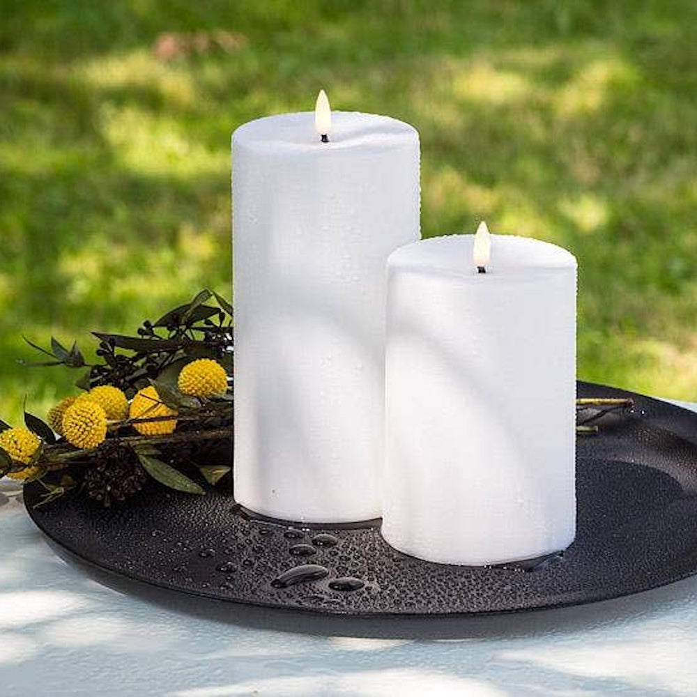 Two white LED pillar candles with water droplets resting on a black tray outdoors.