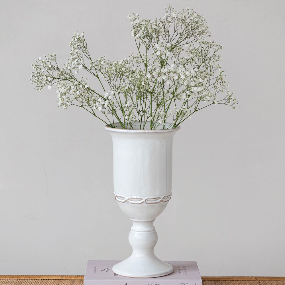 A white pedestal urn vase holding delicate baby's breath flowers, resting on a book against a plain wall.