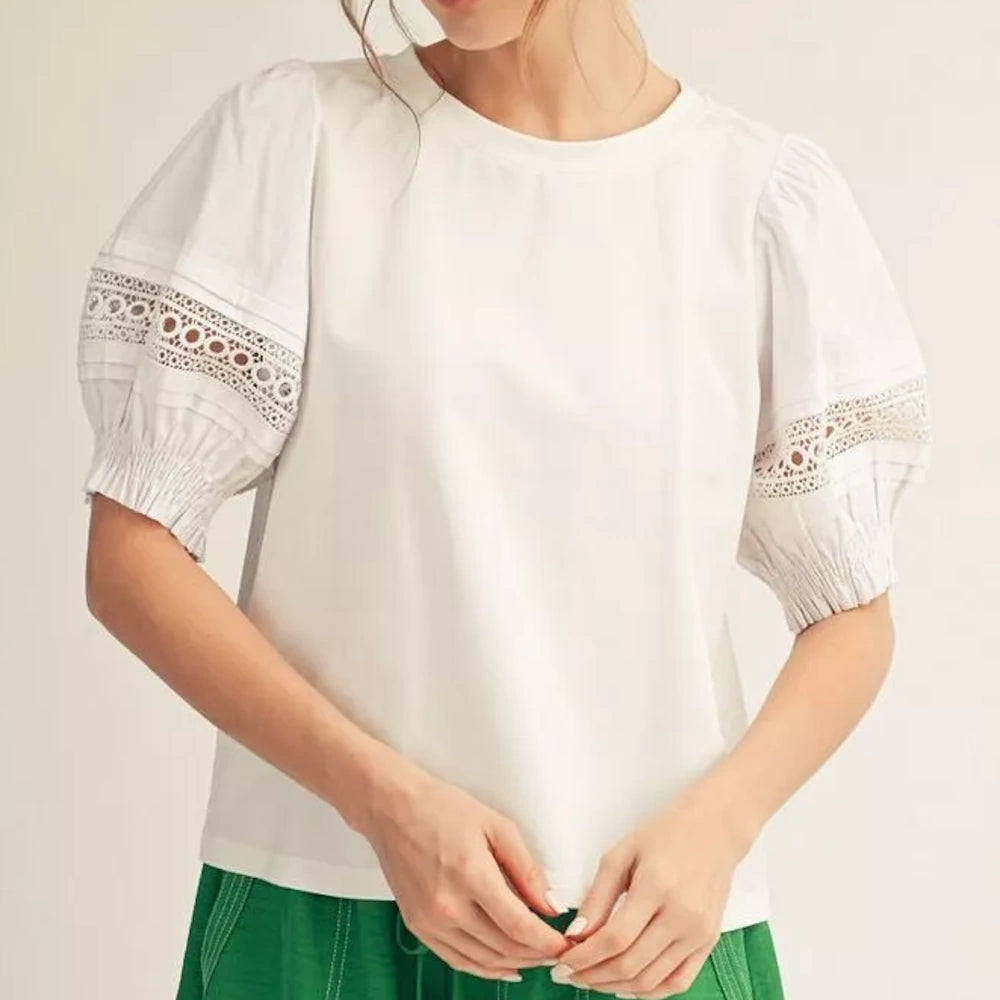 A woman wearing a white blouse with lace-detailed puff sleeves and green pants.