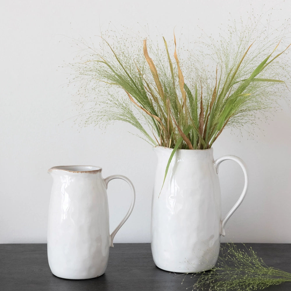 Two white ceramic pitchers of different sizes sit on a dark wood surface, with the larger one holding dried green grass.