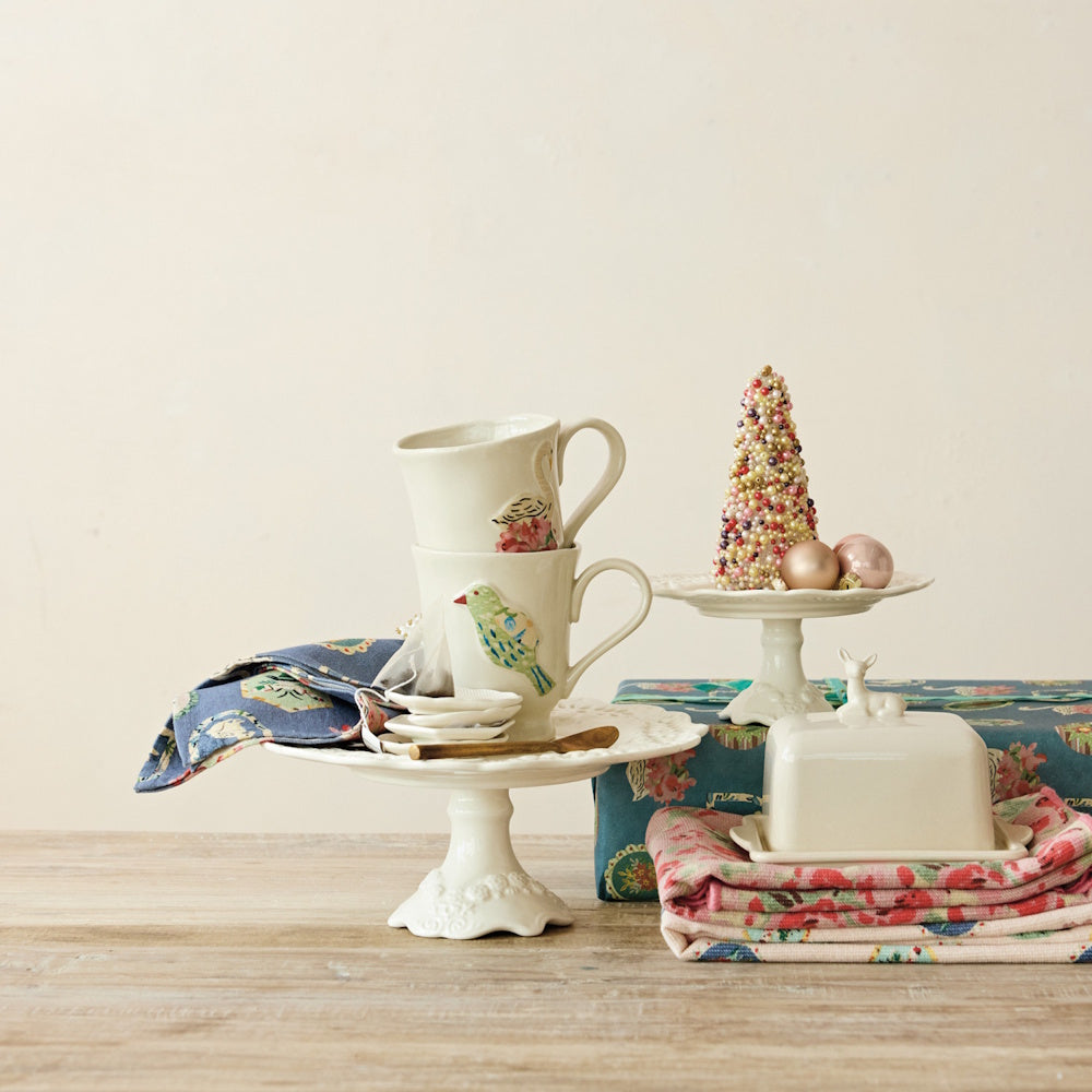 White stoneware pedestals holding decorated mugs and a beaded tree figurine on a table.