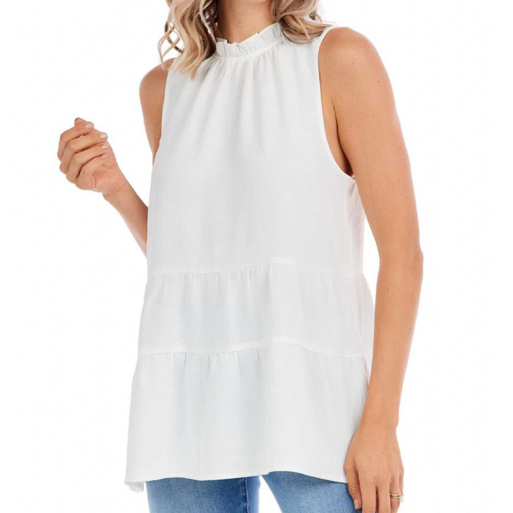A woman wearing a sleeveless white tiered flounce top with a high ruffled neckline, paired with blue jeans.