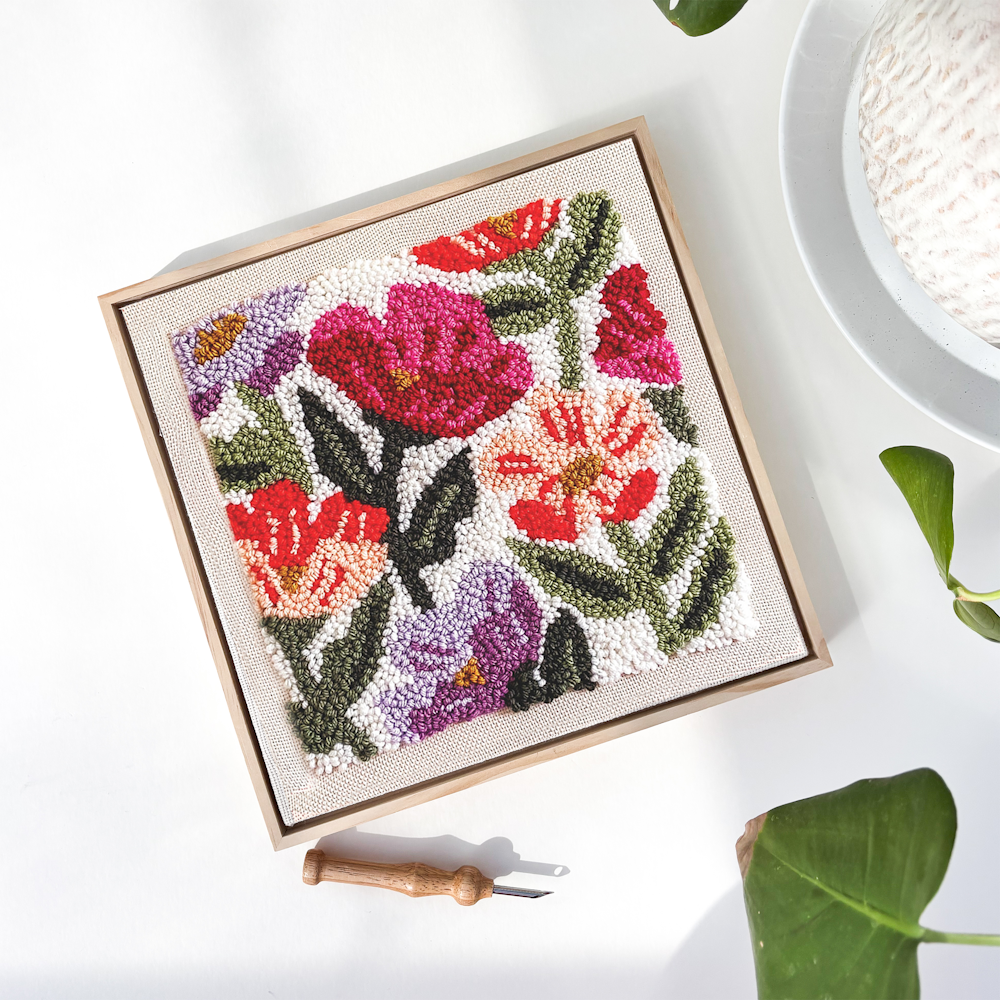 A square wildflower punch needle kit embroidery in a wooden frame with a punch needle tool.