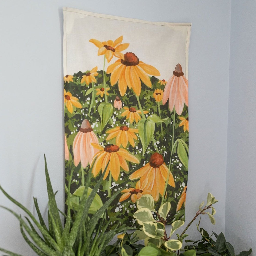 A painted fabric tapestry of yellow and pink coneflowers hanging on a wall above indoor plants.