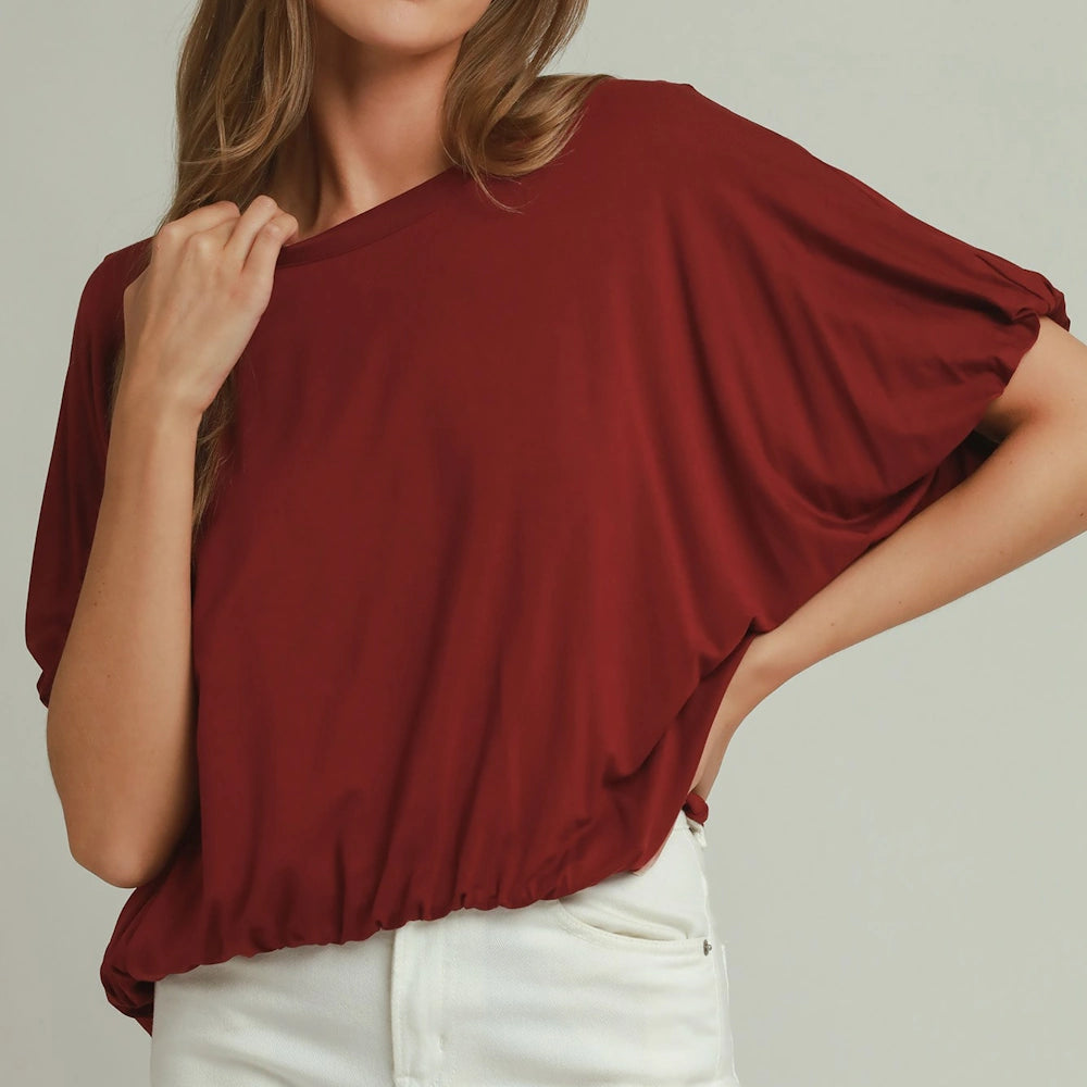 Woman wearing a deep red short-sleeve top with a wide neckline and a cinched bubble hem paired with white pants.
