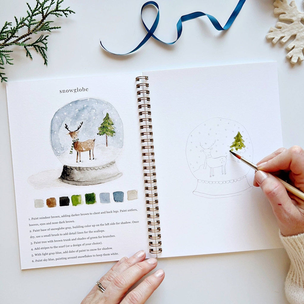 Open watercolor workbook showing step-by-step snow globe painting instructions and line art.