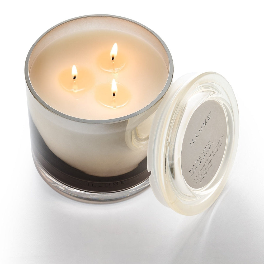A three-wick lit candle in a reflective glass jar with its matching lid beside it.