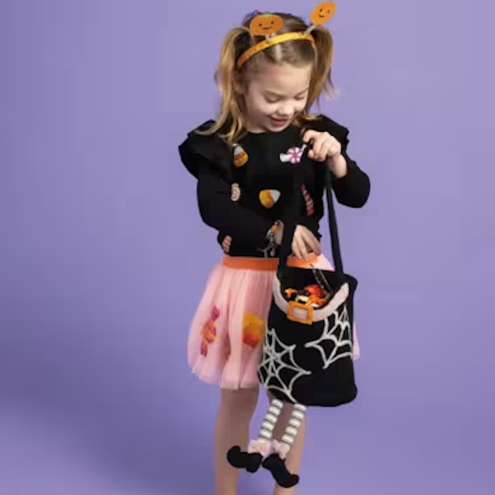 Young girl holding a black witch felt treat bucket with dangling striped legs, wearing a pumpkin headband, black top, and pink skirt.
