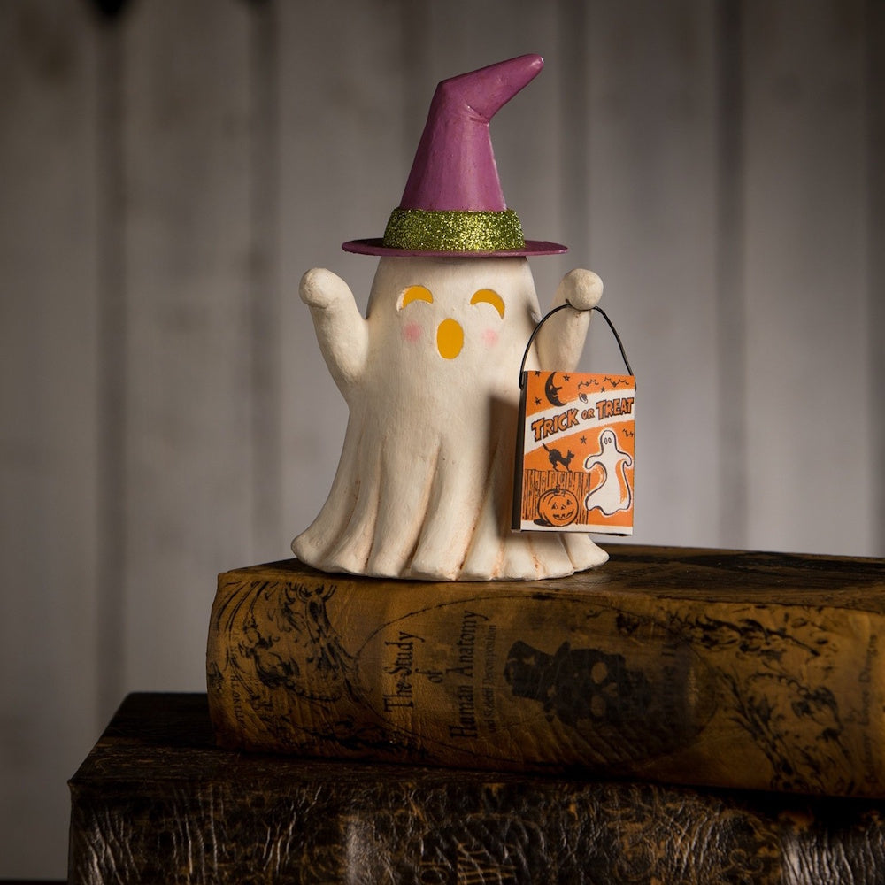 A lit ghost figurine wearing a purple witch hat and holding an orange trick-or-treat bag on an old book.