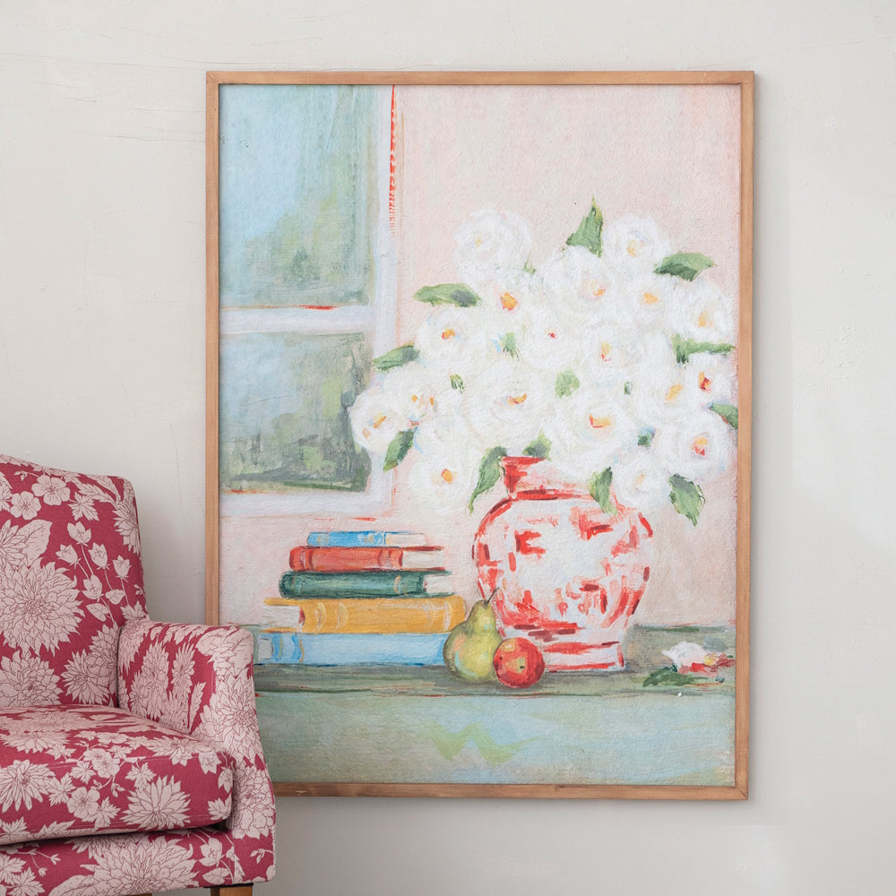 Framed painting of white flowers in a vase and books, next to a red patterned armchair.