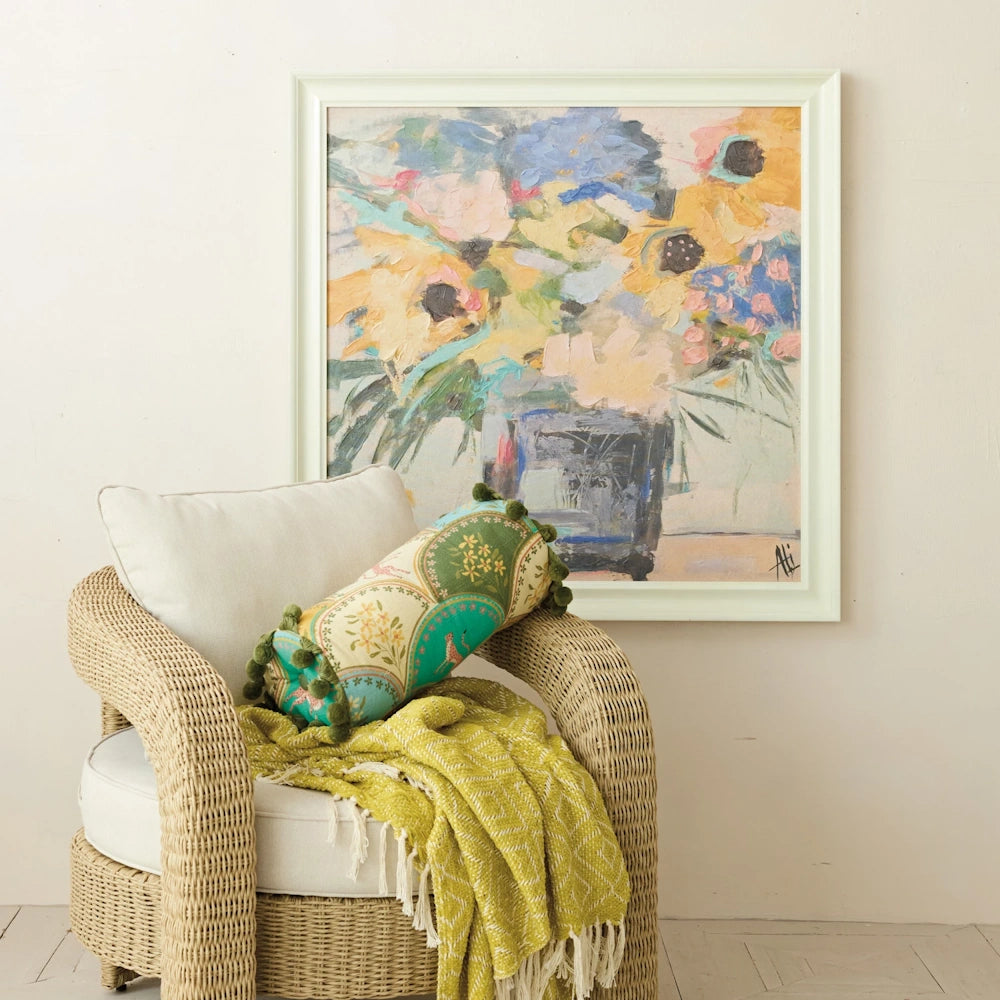 Hand-painted floral canvas wall art hanging above a wicker armchair with a green bolster pillow and yellow throw blanket.