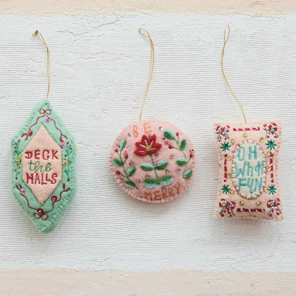 Flat lay view of three assorted pink and green wool felt holiday ornaments against a textured white background.