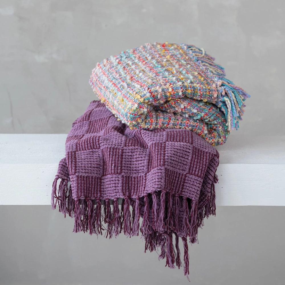 Stacked purple checkered throw blanket and colorful textured knit blanket with fringe on a white shelf.