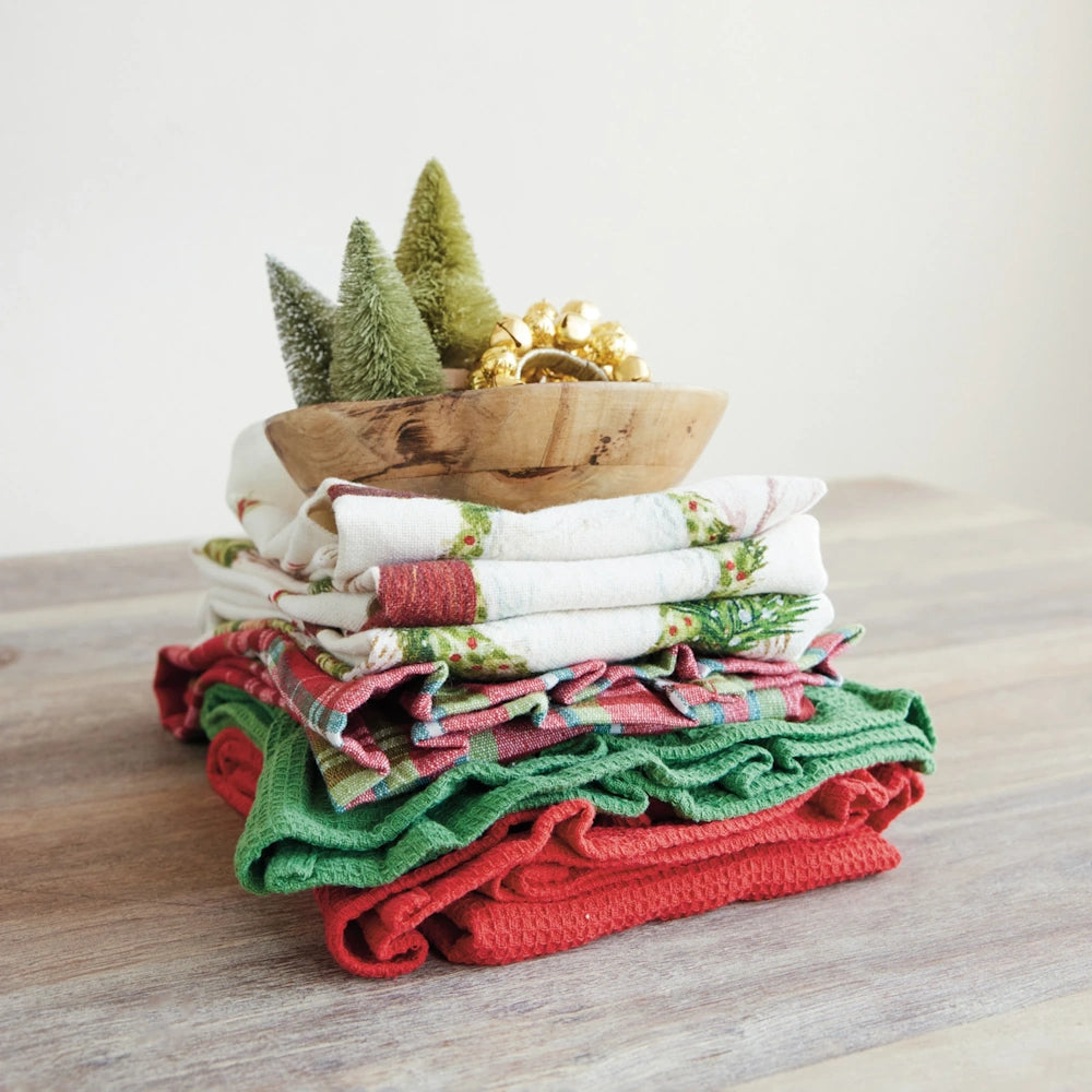 Stack of folded holiday towels topped with a wooden bowl containing mini bottle brush trees and gold bells.