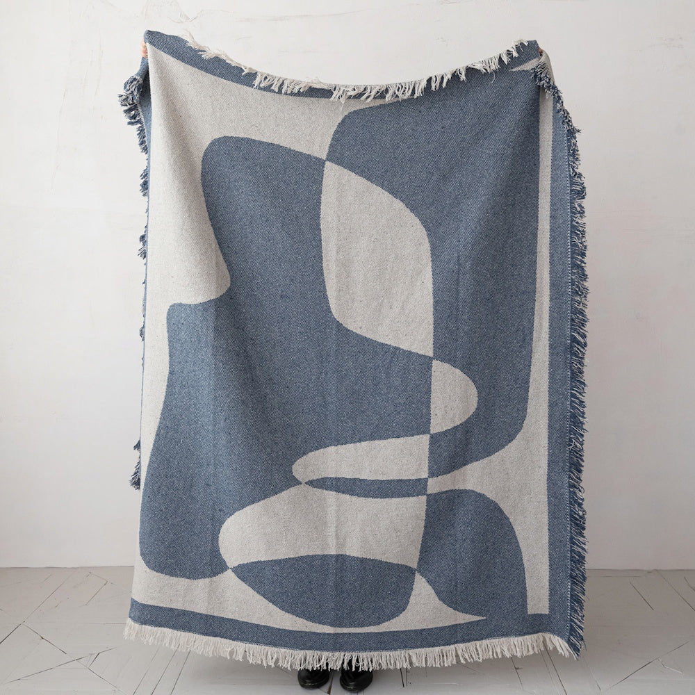 A woven, fringed throw blanket with a large, abstract blue and cream geometric pattern held upright.