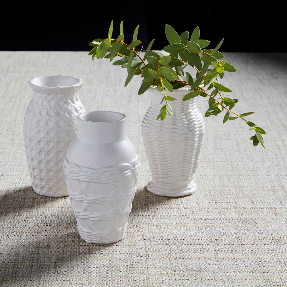 Three white woven stoneware bud vases with green eucalyptus leaves on a textured beige tablecloth.