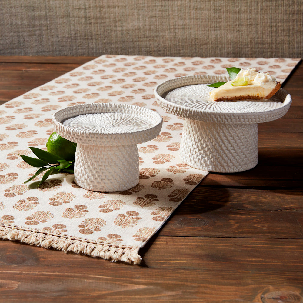 White woven stoneware pedestal servers with a slice of lime pie on a patterned table runner over a wooden surface.