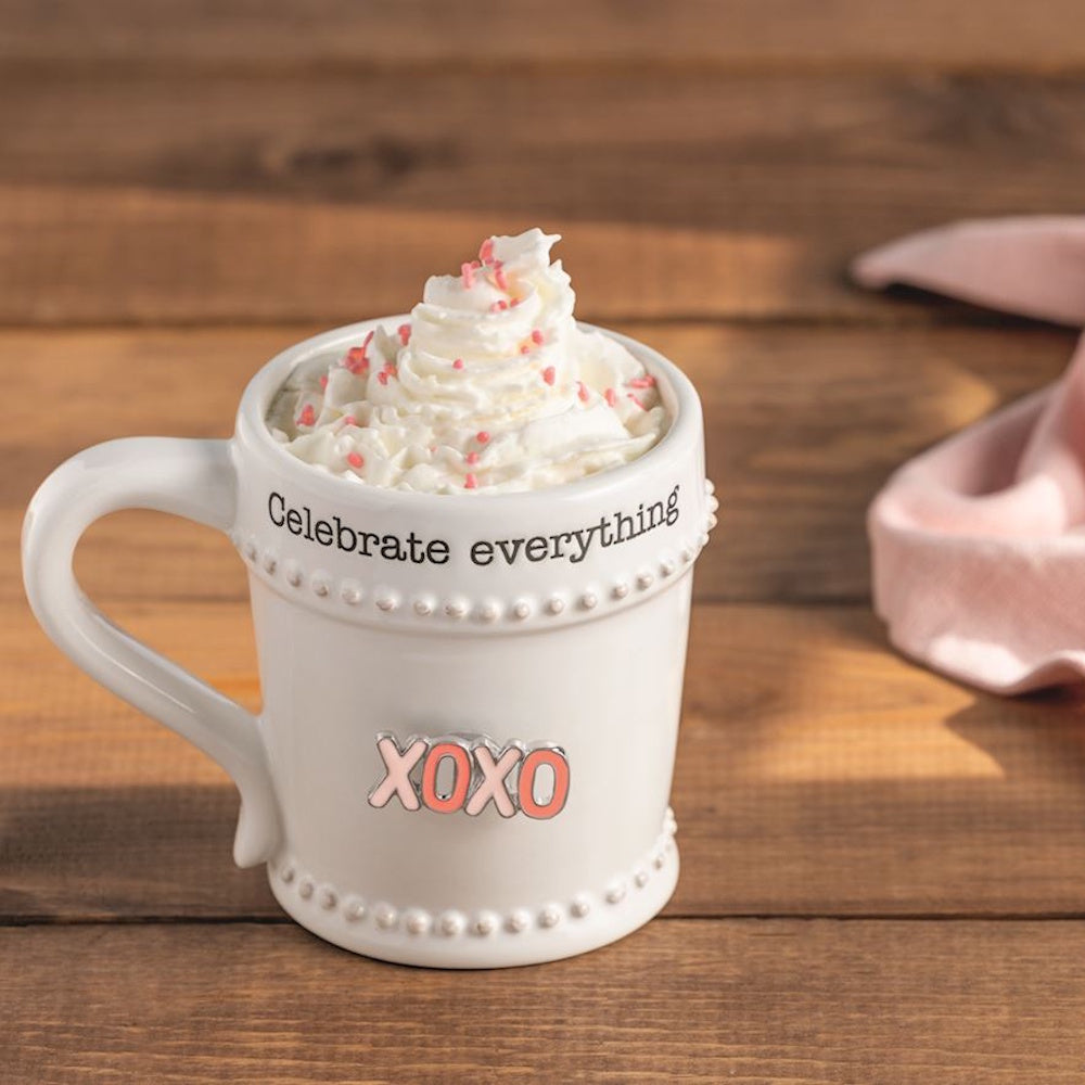 White ceramic mug with "Celebrate everything" text, pink XOXO charm, and whipped cream with sprinkles on a wooden surface.