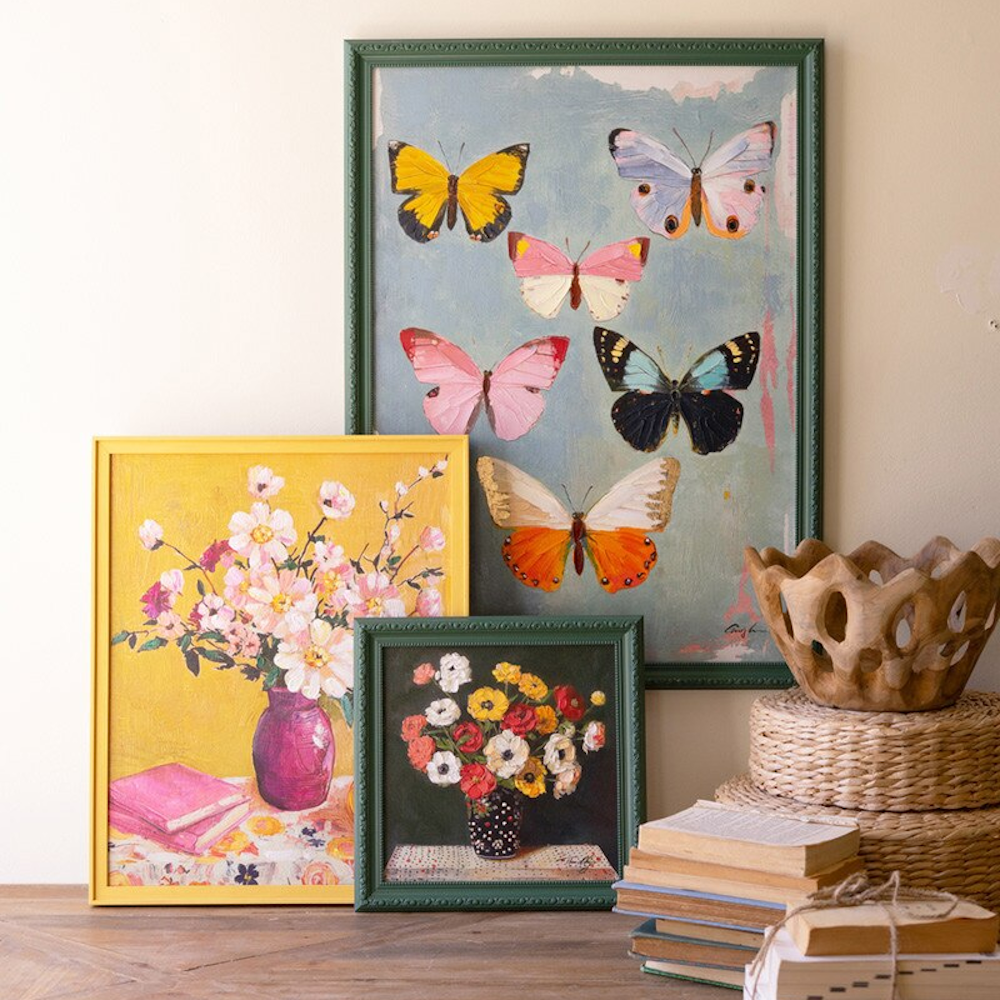 Framed oil paintings of butterflies and flowers arranged on a table with books and a woven basket.