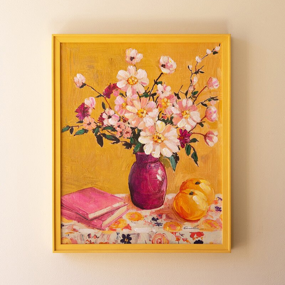 Yellow framed still life oil painting of pink flowers in a purple vase with books and oranges on a yellow background.