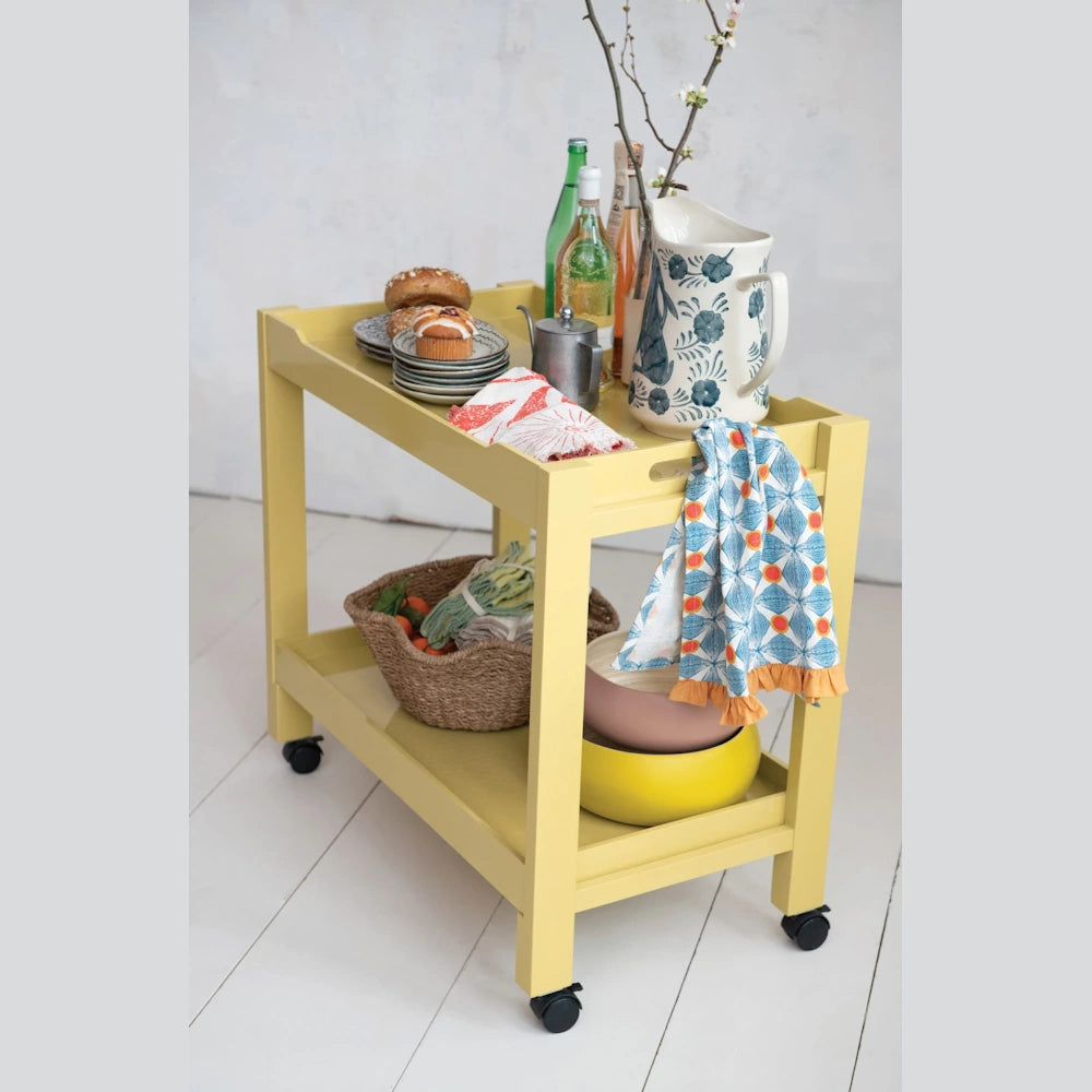 Yellow rolling tea trolley with breakfast items, patterned towels, and drinks on white wooden floors.