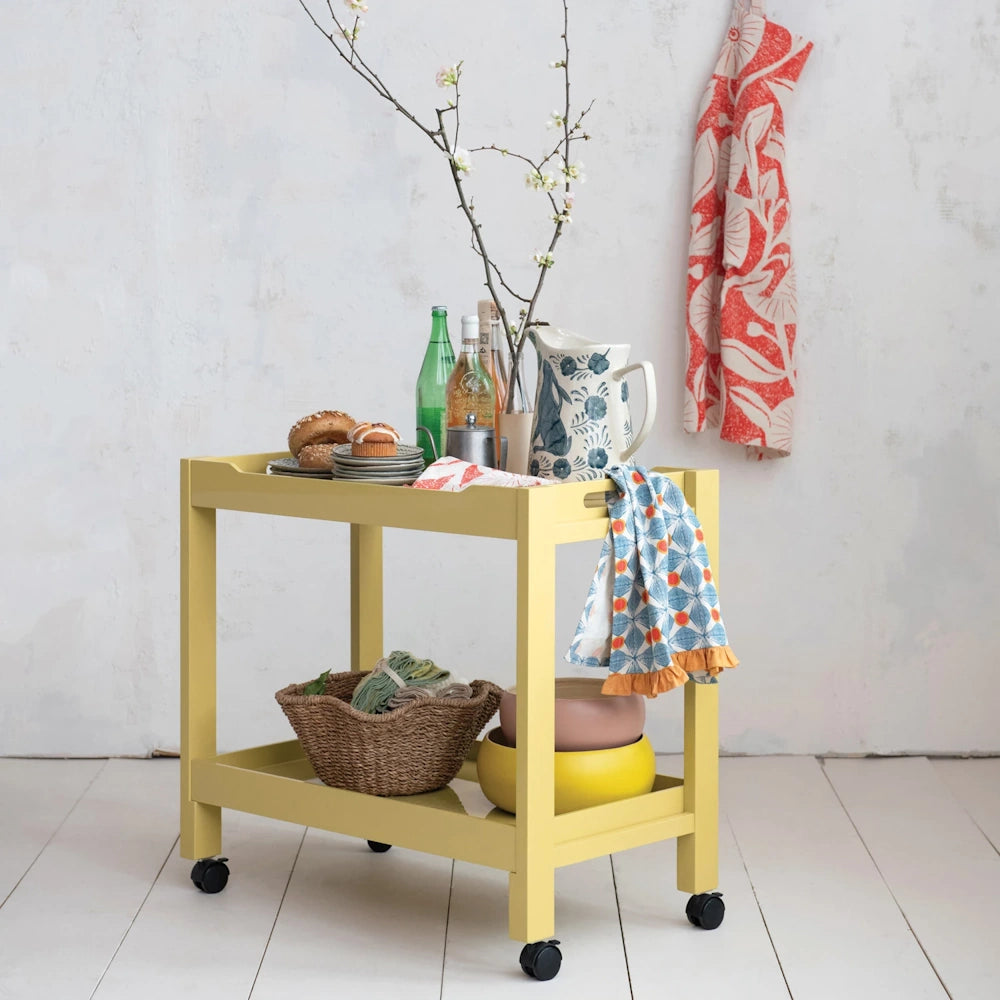 Yellow two-tier rolling bar cart with breakfast treats, pitcher, and woven storage basket.
