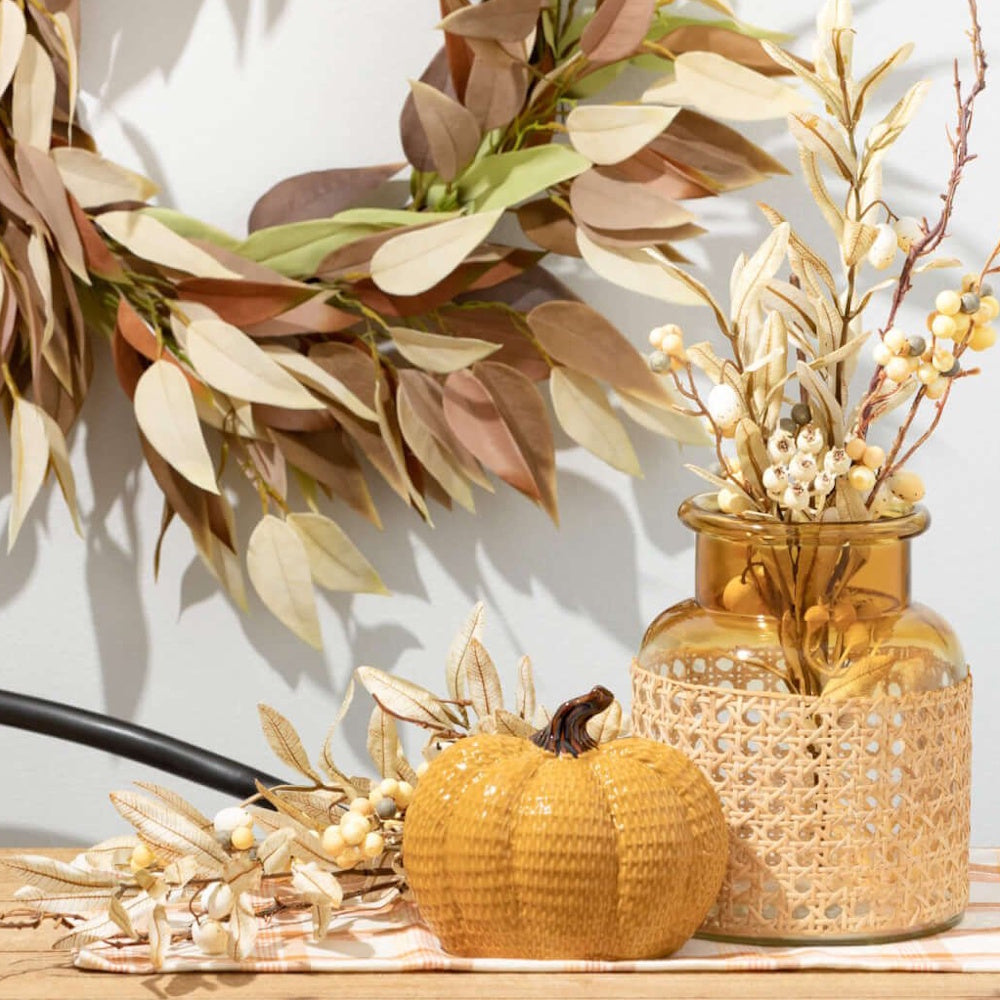 A yellow textured ceramic pumpkin next to an amber glass vase with cream berries and an autumn leaf wreath.
